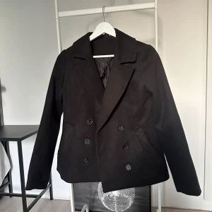 Svart kavaj med dubbelknäppning från Divided - Stilren svart kavaj från Divided med dubbelknäppning och klassisk krage. Jackan har två fickor framtill och är helfodrad. Perfekt för dig som vill ha en tidlös och snygg look.