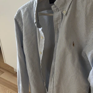 Blåvit randig skjorta från Ralph Lauren - Klassisk blå och vit randig skjorta från Ralph Lauren med knappar framtill och broderad logga på bröstet. Skjortan har lång ärm och button-down krage, perfekt för en stilren look. Perfekt till sommaren. Slim fit. 