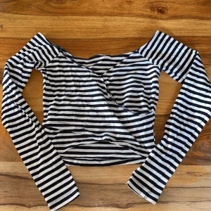 Svartvit randig offshoulder topp från BikBok - Säljer en svartvit randig långärmad topp från BikBok. Modellen är offshoulder med croppad passform och omlott-detalj framtill. Perfekt för dig som gillar en trendig och stilren look.