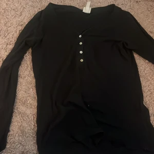 Svart långärmad topp med knappar - Svart långärmad topp med v-ringning och fyra vita knappar framtill. Enkel och stilren design som passar till mycket. Mjuk och skön i materialet.