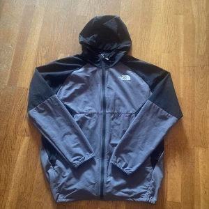 Svart och grå vindjacka från The North Face - Säljer en snygg vindjacka från The North Face i svart och grått. Jackan har huva, dragkedja framtill och två sidofickor. Perfekt för blåsiga dagar och enkel att matcha med det mesta. Logga på bröstet.