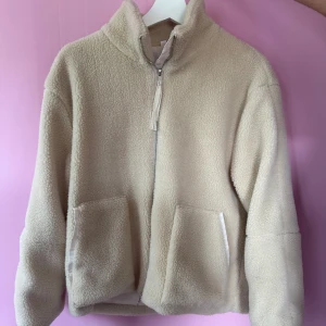 Beige teddyjacka med dragkedja - Mysig beige teddyjacka med hög krage och dragkedja framtill. Jackan har två stora fickor och är perfekt för kyligare dagar. Enkel och stilren design som passar till många outfits.