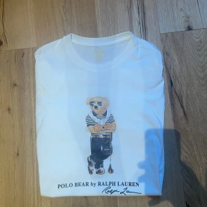 Vit t-shirt med Polo Bear från Ralph Lauren - Säljer en vit t-shirt från Ralph Lauren med det ikoniska Polo Bear-motivet på framsidan. T-shirten har en normal passform och är tillverkad i mjuk bomull. Perfekt för en avslappnad stil med en touch av lyx.
