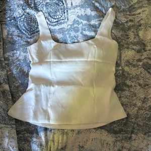 Vit satinlinnetopp med rosettdetalj - Säljer en vit linnetopp i satin med breda axelband och en stor dekorativ rosett baktill. Toppen har en figurnära passform och är perfekt för att lyfta en outfit med sin eleganta look.