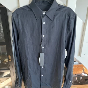 Svart skjorta från J.Lindeberg - Snygg svart skjorta från J.Lindeberg i slim fit-modell. Perfekt för dig som gillar stilrena och enkla plagg. Helt ny, aldrig använd. Skriv gärna vid funderingar. 
