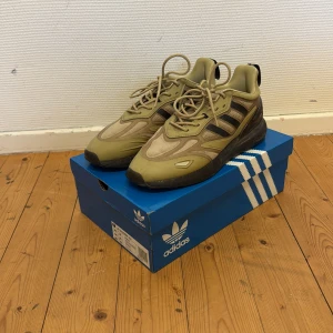 Adidas ZX 2K Boost 2.0 sneakers i beige och svart - Säljer ett par Adidas ZX 2K Boost 2.0 sneakers i beige med svarta detaljer och svart sula. Skorna har snörning och är tillverkade i syntetmaterial med meshpaneler för extra andningsförmåga. Perfekta för dig som gillar stilrena och bekväma sneakers. 44 o 2/3 men sitter som 44 mer! 