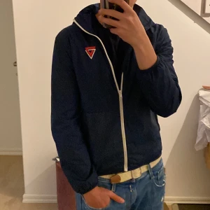 Blå och vit vindjacka med huva - Modellen har vanligtvis storlek M. Snygg blå vindjacka med vita detaljer och huva. Jackan har dragkedja framtill, vit logga på bröstet och vita paneler på sidorna och nedtill. Perfekt för en avslappnad stil. 