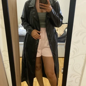 Svart lång skinnkappa med bälte - Säljer en svart lång skinnkappa med klassisk krage och bälte i midjan. Kappan har en rak passform och är perfekt för dig som vill ha en stilren och edgy look. Passar till många olika outfits och ger en cool vibe.