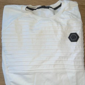 Vit långärmad tröja från Philipp Plein - Säljer en stilren vit långärmad tröja från Philipp Plein med diskreta horisontella sömmar på bröstet och svart logomärke på vänster sida. Tröjan har rund halsringning och är perfekt för en clean look.