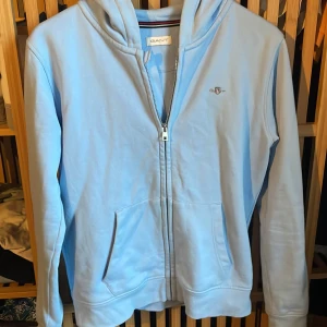 Ljusblå hoodie med dragkedja från GANT - Säljer en ljusblå hoodie från GANT med dragkedja och fickor framtill. Tröjan har huva och ett litet broderat GANT-logo på bröstet.