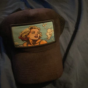 Revenite keps - Snygg Svart revenite keps med nät baktill och justerbar rem. Framtill finns ett färgglatt pop art-tryck med en blond kvinna och bubblor mot blå bakgrund. Perfekt för dig som gillar unika accessoarer med retrostil.