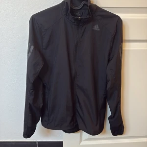 Svart löparjacka från Adidas - Säljer en svart löparjacka från Adidas i herrstorlek XS. Jackan har dragkedja framtill, lång ärm och diskreta reflexdetaljer på ärmarna. Perfekt för löpning eller träning utomhus. Adidas-logga på bröstet.
