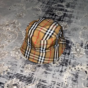 Rutig bucket hat från Burberry - Säljer en klassisk bucket hat från Burberry i deras ikoniska beigea rutiga mönster med svarta, vita och röda detaljer. Hatten är tillverkad i bomull och har en mjuk, rund form. Perfekt accessoar för att lyfta din stil.