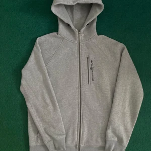 Grå hoodie från Sail Racing - Säljer en grå hoodie från Sail Racing med dragkedja framtill och en extra bröstficka med dragkedja. Tröjan har huva och broderad logga på bröstet samt tryck på ryggen. Perfekt för en avslappnad stil. Ett litet defekt på dragkedjan men den fungerar!