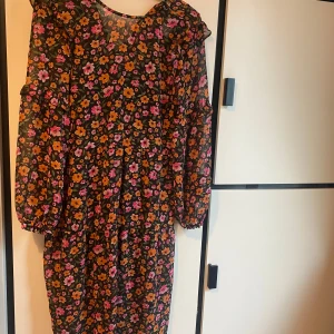 Blommig klänning från H&M Divided - Supersöt blommig klänning från H&M Divided i svart med orange och rosa blommor. Klänningen har långa ärmar med lätt puff och volangdetaljer upptill. Perfekt för dig som gillar färg och mönster!