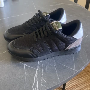 Svarta Valentino rockrunners  - Säljer ett par svarta Valentino sneakers med vita hälpartier och grå mockadetaljer. Skorna har snörning, rund tå och platt sula. Märket syns tydligt på sulan. Perfekta för dig som gillar stilrena och exklusiva sneakers. Vet ej om de e äkta bra skick 4O men passar 41