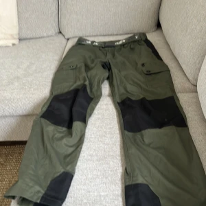 Gröna friluftsbyxor från Pinewood - Säljer ett par gröna friluftsbyxor från Pinewood Outdoor Collection med svarta förstärkningar på knän och benslut. Byxorna har flera fickor och medföljer ett kamouflagemönstrat bälte med texten 'DENIM'. Perfekta för utomhusaktiviteter.