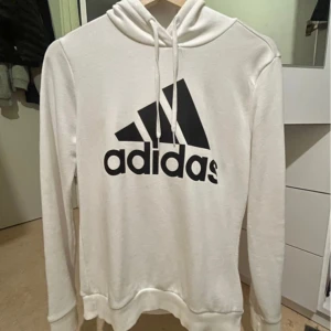 Vit hoodie från Adidas - Vit hoodie från Adidas med stor svart logga framtill. Tröjan har huva med dragsko och långa ärmar. Perfekt för en sportig och avslappnad stil.