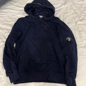 Mörkblå hoodie från C.P. Company - Säljer en mörkblå hoodie från C.P. Company med klassisk huva och magficka. Tröjan har en ikonisk linsdetalj på ärmen och ribbade muddar. Perfekt för en avslappnad och stilren look. Storlek m men passar som S
