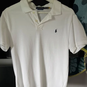 Vit pikétröja från Polo Ralph Lauren - Klassisk vit pikétröja från Polo Ralph Lauren med korta ärmar och broderad logga på bröstet. Slim fit-modell med krage och knappar framtill. Perfekt för en stilren och tidlös look. Storlek xl i barnstorlek vilket motsvarar s i vanlig storlek!