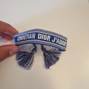 Blått armband från Christian Dior J'Adior - Helt nytt armband!! Säljer ett blått tygarmband från Christian Dior J'Adior med vit text och dekorativa tofsar i ändarna. Armbandet har ett flätat knytband och är perfekt för att ge din outfit en exklusiv touch.