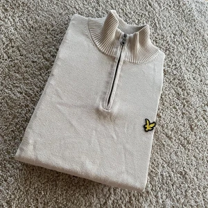 Beige stickad halvzip-tröja från Lyle & Scott - Snygg beige stickad tröja från Lyle & Scott med halv dragkedja och ribbad krage. Tröjan har det klassiska gula fågel-loggan broderad på bröstet. Perfekt för lager-på-lager och stilrena outfits.