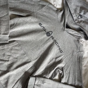 Grå hoodie från Sail Racing - Säljer en grå hoodie från Sail Racing med svart logga på bröstet. Tröjan har huva och en stor magficka framtill. Perfekt för en avslappnad stil och enkel att matcha med det mesta.