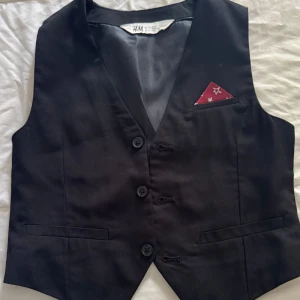 Svart kostymväst från H&M - Stilren svart kostymväst från H&M med tre knappar framtill och två fickor. Västen har en klassisk krage och ett dekorativt rött näsdukstyg med stjärnor i bröstfickan. Perfekt för att lyfta din outfit( till barn 8-9 år.)