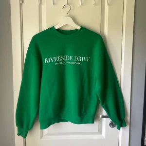 Grön sweatshirt  - Säljer en grön sweatshirt med vit text 'Riverside Drive Upper East Side, New York' på framsidan. 