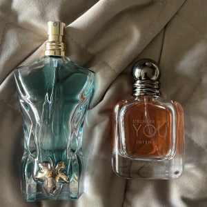 Jean Paul Gaultier Le Male och Emporio Armani Stronger With You Intensely herrparfymer - Söker Byte Två herrparfymer: Le Male från Jean Paul Gaultier i en blå, kroppformad flaska med gulddetaljer och Stronger With You Intensely från Emporio Armani i en kompakt, rundad glasflaska med silverkork och bärnstensfärgad vätska. 