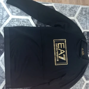 Svart sweatshirt från Emporio Armani EA7 - Säljer en svart långärmad sweatshirt från Emporio Armani EA7 med stort guldigt logotryck på bröstet. Tröjan har rund halsringning och klassisk passform. Perfekt för dig som gillar stilrena märkeskläder.