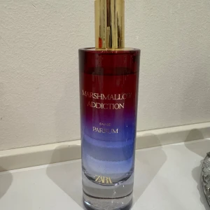 Marshmallow Addiction Parfym Zara - En parfymflaska med guldlock och en snygg färgövergång från rött till blått. Flaskan har texten 'Marshmallow Addiction' i guld och rymmer en söt, gourmanddoft. Perfekt för dig som gillar unika och lekfulla dofter.