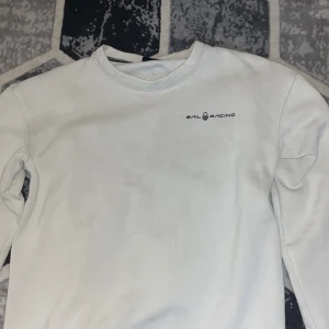 Vit sweatshirt från Sail Racing - Vit sweatshirt från Sail Racing med rund hals och diskret logga på bröstet. Tröjan är tillverkad i en mjuk bomulls- och polyestermix och har långa ärmar. Perfekt för en clean och stilren look.