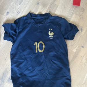 Frankrike fotbollströja Mbappé #10 - Mörkblå fotbollströja från Frankrikes landslag med guldiga detaljer. Nummer 10 och Mbappé på ryggen, samt landslagets emblem med två stjärnor på bröstet. Kortärmad modell i lätt och sportigt material.