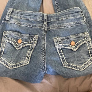Blå bootcut jeans från Gina Tricot - Säljer ett par klassiska blå jeans från Gina Tricot i bootcut-modell. Jeansen har dekorativa bakfickor med kontrastsömmar och knappdetaljer. Perfekta för dig som gillar en lite utsvängd passform.