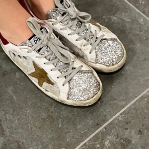 Glittriga sneakers från Golden Goose - Säljer ett par vita sneakers från Golden Goose med silvriga glitterdetaljer på tån och tungorna, guldstjärna på sidan och röda hälkappor. Skorna har snörning och en klassisk rund tå. Perfekta för dig som vill sticka ut med lite extra bling!