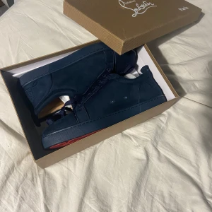 Mörkblå sneakers från Christian Louboutin - Säljer ett par mörkblå sneakers från Christian Louboutin med klassisk röd sula och snörning. Skorna har en stilren design i mocka och är perfekta för dig som vill sticka ut med exklusiva detaljer.