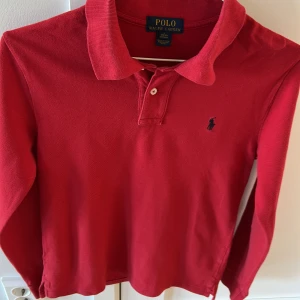 Röd pikétröja från Polo Ralph Lauren - Säljer en klassisk röd pikétröja från Polo Ralph Lauren med lång ärm och krage. Tröjan har knappar vid halsen och den ikoniska broderade loggan på bröstet. Perfekt för en stilren och tidlös look.