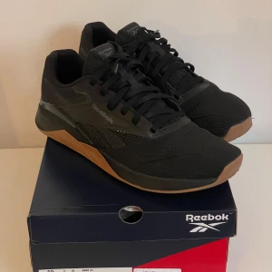 Träningsskor  Reebok Nano X4 - Säljer ett par svarta Reebok Nano X4 skor med bruna gummisulor och snörning. Skorna har meshdetaljer för extra andningsförmåga och en stabil platt sula som passar perfekt för Crossfit. 