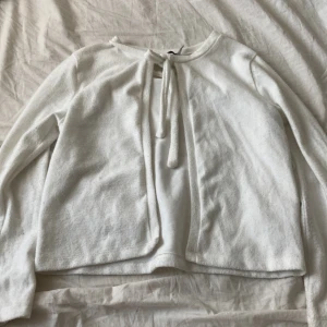 Vit kofta med knyt framtill - Säljer en söt vit kofta med knyt framtill. Tröjan har långa ärmar och är gjord i ett mjukt material som känns skönt mot huden. Perfekt att slänga över en topp för en enkel och stilren look.