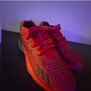 Röda Adidas basket sneakers med svarta detaljer - Säljer ett par röda Adidas sneakers med svarta detaljer och snörning. Skorna har en sportig design med stickade mönster på ovandelen och platt sula. Perfekta för dig som gillar färgstarka och unika sneakers.