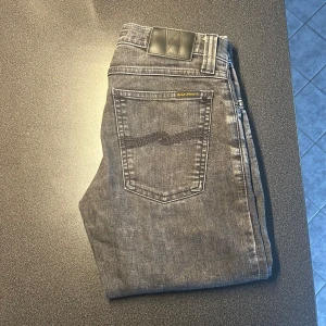Grå  jeans från Nudie Jeans - Säljer ett par grå skinny jeans från Nudie Jeans med klassisk femficksdesign och svarta detaljer. Jeansen har en smal passform och är tillverkade i stretchigt denim för extra komfort.