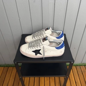 Golden goose - Snygga vita sneakers från Golden Goose med svart stjärna på sidan och blå hälkappa. Skorna har grå snörning och klassisk låg profil. Tillverkade i skinn med coola detaljer och text på sidan. Perfekta för dig som gillar streetstyle och unika skor.