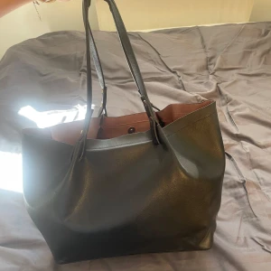 Svart shoppingväska i skinn - Stilren svart shoppingväska i skinn. Väskan har långa handtag som gör den enkel att bära på axeln och en rymlig insida med knappstängning upptill. Perfekt för dig som vill ha plats för allt du behöver under dagen.