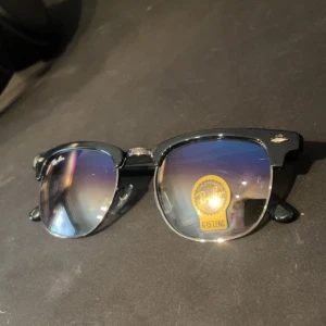 Ray-Ban Clubmaster g15 lens - Snygga Ray-Ban Clubmaster solglasögon med svart båge och silvriga detaljer. Glasen har en mörk ton med blå-lila spegeleffekt och klassisk G-15 lins.