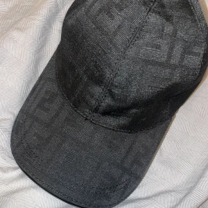 Fendi keps  - Snygg svart keps från Fendi med diskret, invävt logomönster över hela kepsen. Justerbar rem baktill i svart skinn med Fendi-logga. Tillverkad i bomull och detaljer i skinn. Perfekt accessoar för en clean och lyxig look.