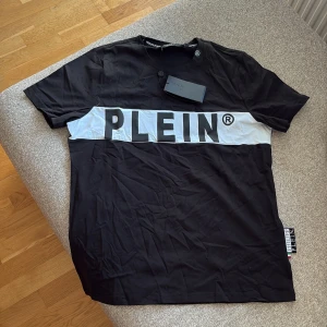 Svart t-shirt från Philipp Plein - Svart t-shirt från Philipp Plein i bomull med vit panel och stor PLEIN-logga över bröstet. Klassisk rund hals och korta ärmar. På ryggen finns en snygg PP-logga i svart och silver. Perfekt för dig som gillar streetwear med lyxig vibe.