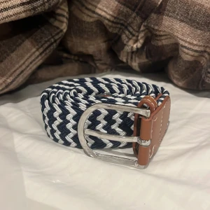 Randigt flätat bälte med skinn-detaljer - Snyggt flätat bälte i marinblått och vitt med bruna skinn-detaljer och silverspänne. Bältet har ett randigt mönster och är perfekt för att ge outfiten en fräsch touch. Passar till jeans eller chinos och har en flexibel, elastisk känsla.