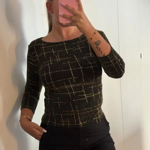 Glittrig svart topp från Xanaka - Svart glittrig topp från Xanaka med guldiga rutmönster och 3/4-ärm. Sitter figurnära och har en rundad halsringning. Perfekt för dig som vill sticka ut med lite extra glitter och coolt mönster.