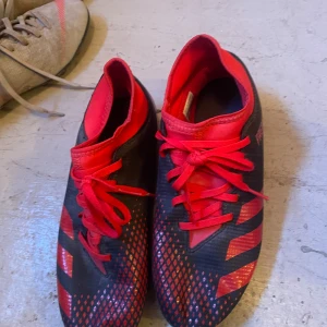 Adidas Predator fotbollsskor röd/svart - Snygga Adidas Predator fotbollsskor i svart och rött med coola mönster och röda snören. Skorna har en låg profil och är tillverkade i syntetmaterial med greppig sula för bästa fäste på planen. Perfekta för dig som vill sticka ut på matchen!